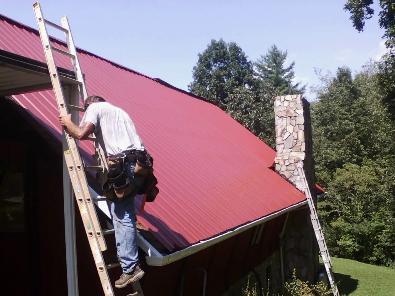 Roofer on a ladder beside a red metal roof and stone chimney — Soffit & Fascia Repair by Roofing Brothers in Happy Valley